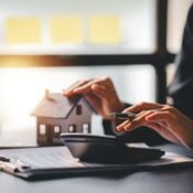 Person sitting at a desk calculates mortgage costs using a calculator while holding a small model house. A laptop and clipboard with documents are nearby, symbolizing cashout refinancing.