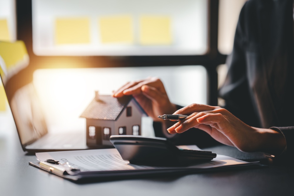 Person sitting at a desk calculates mortgage costs using a calculator while holding a small model house. A laptop and clipboard with documents are nearby, symbolizing cashout refinancing.