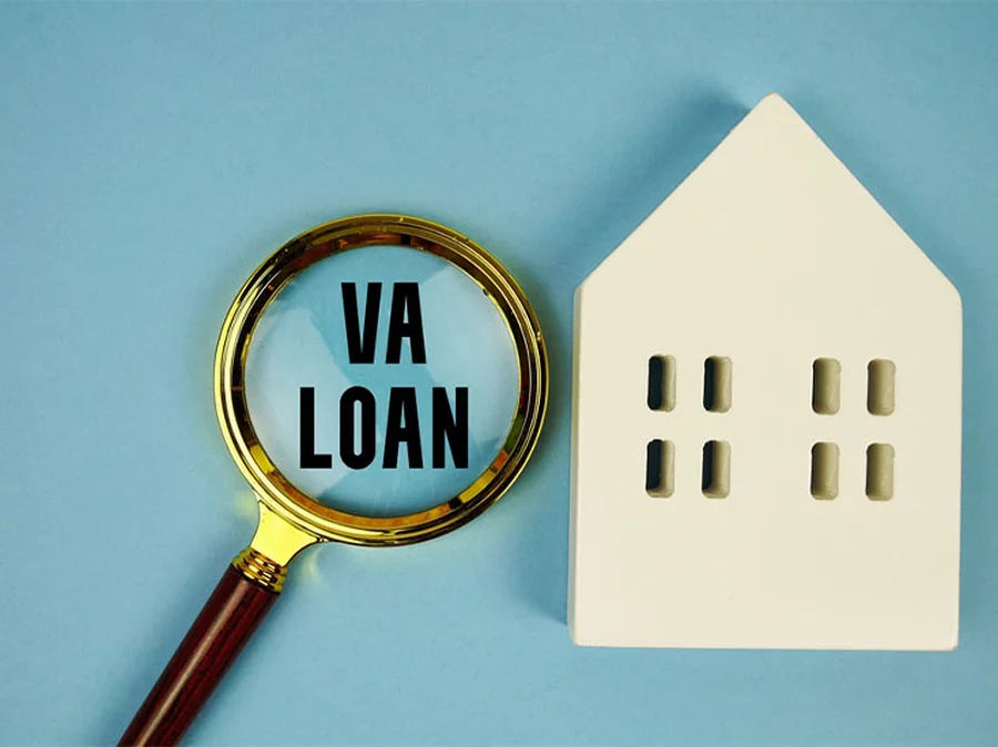 “VA loans” shown in a magnifying glass placed next to a cream-colored wooden 2D home.